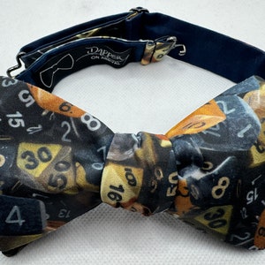 May include: A black bow tie with a pattern of gold and black numbers. The bow tie has an adjustable strap with a metal clasp. The text "DAPPER ON ARRIVAL" is printed on the strap.