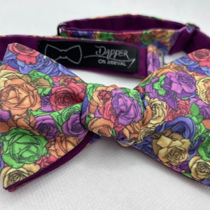 May include: A purple bow tie with a floral pattern featuring a variety of colorful roses. The bow tie has a purple backing and a black label that reads "Dapper On Arrival".