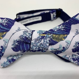 May include: A white bow tie with a blue and white print of the Great Wave off Kanagawa by Katsushika Hokusai. The bow tie has a navy blue adjustable strap with the text "Dapper On Arrival" printed on it.