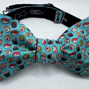 May include: A turquoise bow tie featuring a repeating pattern of various sushi rolls. The bow tie has a black adjustable neckband and the text "Dapper on Arrival" printed on the inside.