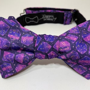 May include: A purple bow tie with a galaxy-themed print of dice with gold numbers. The bow tie has a black adjustable strap.