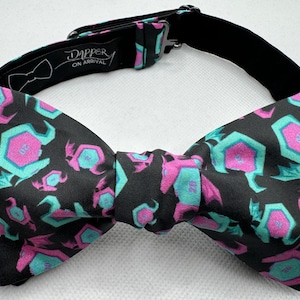 May include: A black bow tie with a repeating pattern of pink and teal dice. The bow tie has an adjustable strap with the text "Dapper On Arrival" printed on it.