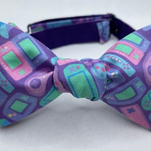 May include: A purple bow tie with a repeating pattern of retro video game consoles in shades of green, pink, and blue. The bow tie has a dark purple adjustable neckband.