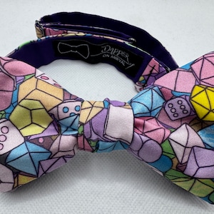May include: A colorful bow tie with a geometric pattern of dice and gemstones. The bow tie is made of a soft, silky fabric and has a purple adjustable strap.