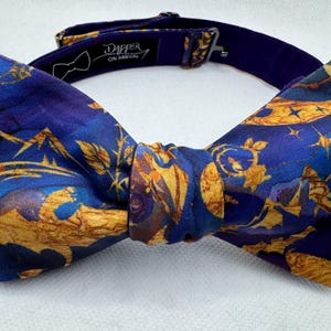 May include: A patterned bow tie in shades of blue and gold, with a dark blue adjustable neckband. The bow tie displays a repeating pattern of gold shapes and stars against a blue backdrop. The text "Dapper On Arrival" is visible on the neckband.