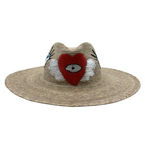 Könnte beinhalten: Breitkrempiger Strohhut mit gesticktem Herzdesign. Das Herz ist rot mit einem weiß umrandeten Auge und weißen Flügeln. Der Hut hat eine natürliche beige Farbe und ist für den Sonnenschutz geeignet.