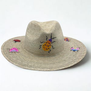 May include: A beige straw fedora hat with a wide brim, adorned with embroidered colorful insect designs. The insects are in various colors, including red, pink, yellow, and blue. The hat has a classic, structured shape.