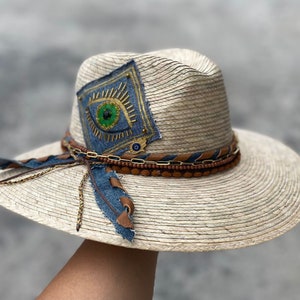 May include: A straw hat with a wide brim, featuring a blue denim patch with a gold eye design and a brown leather band with gold chain accents.