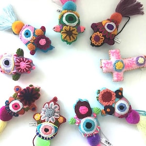 May include: Nine colorful crocheted hand charms with tassels. Each charm features an embroidered eye and floral designs in shades of pink, blue, green, yellow, and red.