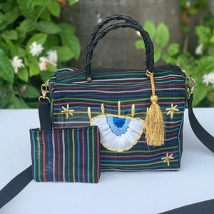 以下が含まれることがあります： 黒と虹色のストライプのメッシュトートバッグ。大きな青い刺繍の目と金のタッセルが付いています。バッグには黒いショルダーストラップと、同じデザインの小さなポーチが付いています。