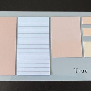 May include: A light gray notepad with a spiral binding on the left side. The notepad features several rectangular and square sticky notes in shades of peach, cream, and white. The text "True Legends" is printed on the bottom right.