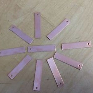 May include: Ten rectangular copper blanks with a hole in the top. The blanks are arranged in a circle on a wooden surface.