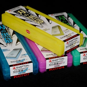 May include: A collection of Shapton ceramic whetstones in various colors, including blue, yellow, pink, and green. Each whetstone is in a plastic case with Japanese text and product information. The cases are labeled with different grit numbers.