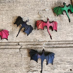 May include: Dragon-shaped earrings in various colors, including green, red, and blue, with silver-colored ear wires. The earrings are displayed on a weathered wooden surface, showcasing their detailed design and craftsmanship.
