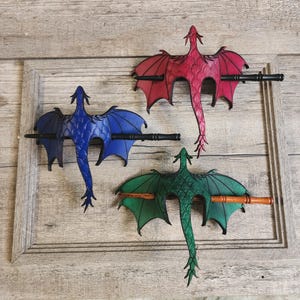 May include: Three dragon-shaped hair barrettes in blue, red, and green. Each dragon has outstretched wings and a long tail, with a black or brown barrette clasping the wings. The barrettes are displayed on a wooden surface.