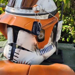 May include: A detailed, close-up view of a Star Wars Clone Trooper helmet. The helmet is primarily white and orange, with black and blue accents. It features a visor, a comms device, and dog tags. The helmet is a replica or prop.