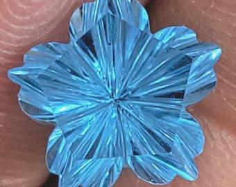 May include: A faceted, light blue gemstone shaped like a five-petal flower. The gemstone has a starburst pattern radiating from the center. The facets catch the light, creating a sparkling effect. The gemstone is held in a hand.