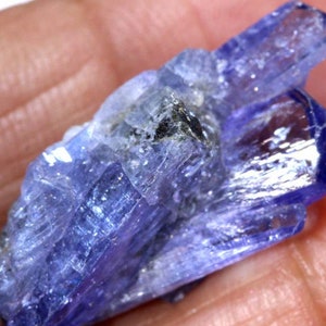 May include: A cluster of blue-purple, translucent, faceted, elongated crystals. The crystals are arranged in a cluster, with some crystals overlapping others. The crystals have a glassy luster.