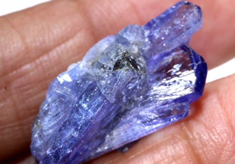 May include: A cluster of blue-purple, translucent, faceted, elongated crystals. The crystals are arranged in a cluster, with some crystals overlapping others. The crystals have a glassy luster.