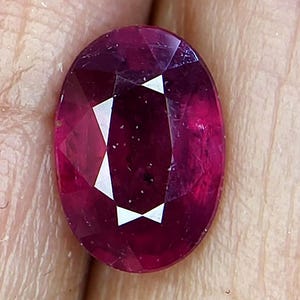 May include: A faceted, oval-shaped ruby gemstone. The gem is a deep, rich red color with white reflective facets. The stone is held in a hand, showcasing its polished surface and clarity. This gemstone is suitable for jewelry.