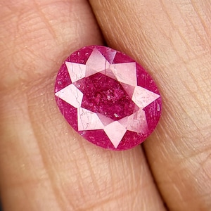 May include: A deep red oval-shaped gemstone with a star-shaped cut. The gemstone is faceted and has a high polish.