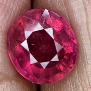 Può includere: Una gemma di rubino sfaccettata di forma ovale. Il rubino è di un rosso intenso e vibrante con sfaccettature geometriche bianche al centro. La gemma è tenuta in una mano, con il tono della pelle che fornisce uno sfondo contrastante.