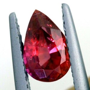 Puede incluir: Una piedra preciosa facetada en forma de lágrima con un tono rojo intenso. La piedra preciosa está sujeta por un par de pinzas metálicas.