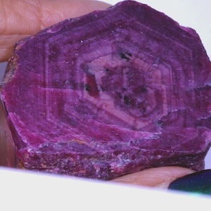 Peut inclure: Une pierre précieuse de rubis brut, d'un magenta profond, avec un motif hexagonal en son centre. La pierre présente un aspect stratifié avec des stries visibles et une texture légèrement rugueuse. Le rubis est tenu dans une main, révélant son état naturel et non poli.