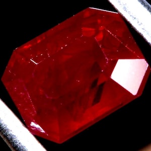 May include: A deep red gemstone with a faceted cut. The gemstone is a rectangular shape with a slightly rounded edge. The gemstone is set in a silver setting.