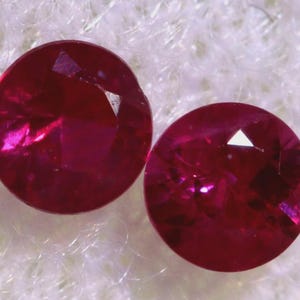 May include: Two round, faceted ruby gemstones with a deep red colour. The gemstones are set against a white, fuzzy background.