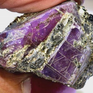 May include: A raw gemstone with a deep purple colour, displaying natural crystalline structures. The stone is partially encased in a matrix of dark grey and gold-coloured minerals, highlighting its rough, unpolished texture. The gemstone is held by a person.