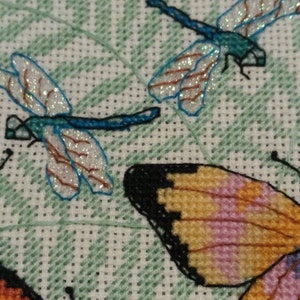 K&ouml;nnte beinhalten: Kreuzstich-Stickerei mit zwei blauen Libellen mit glitzernden Fl&uuml;geln und einem Schmetterling mit orangefarbenen, rosa und schwarzen Fl&uuml;geln auf einem gr&uuml;nen Farn-Hintergrund.