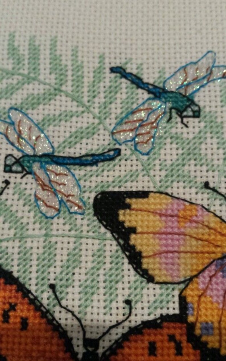 K&ouml;nnte beinhalten: Kreuzstich-Stickerei mit zwei blauen Libellen mit glitzernden Fl&uuml;geln und einem Schmetterling mit orangefarbenen, rosa und schwarzen Fl&uuml;geln auf einem gr&uuml;nen Farn-Hintergrund.