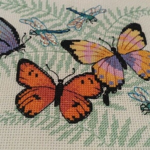 K&ouml;nnte beinhalten: Eine Kreuzstich-Stickerei mit vier Schmetterlingen und Libellen. Die Schmetterlinge sind in den Farben Lila, Orange und Gelb gehalten, w&auml;hrend die Libellen blau und gr&uuml;n sind.
