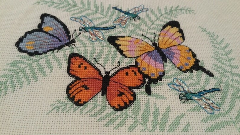 K&ouml;nnte beinhalten: Eine Kreuzstich-Stickerei mit vier Schmetterlingen und Libellen. Die Schmetterlinge sind in den Farben Lila, Orange und Gelb gehalten, w&auml;hrend die Libellen blau und gr&uuml;n sind.