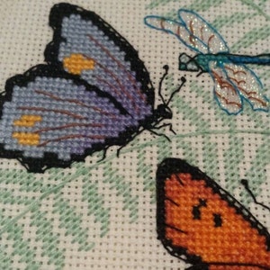 K&ouml;nnte beinhalten: Kreuzstich-Stickerei mit drei Schmetterlingen. Ein lilafarbener Schmetterling mit gelben Akzenten, ein orangefarbener Schmetterling und eine blaue Libelle.
