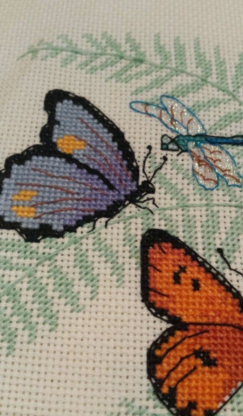 K&ouml;nnte beinhalten: Kreuzstich-Stickerei mit drei Schmetterlingen. Ein lilafarbener Schmetterling mit gelben Akzenten, ein orangefarbener Schmetterling und eine blaue Libelle.