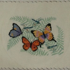 K&ouml;nnte beinhalten: Ein wei&szlig;er Stoff mit einem gr&uuml;nen Farnmuster und gestickten Schmetterlingen. Die Schmetterlinge sind lila, orange und rosa mit blauen Libellenakzenten.