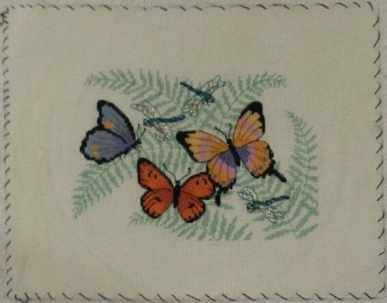 K&ouml;nnte beinhalten: Ein wei&szlig;er Stoff mit einem gr&uuml;nen Farnmuster und gestickten Schmetterlingen. Die Schmetterlinge sind lila, orange und rosa mit blauen Libellenakzenten.