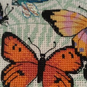K&ouml;nnte beinhalten: Kreuzstich-Stickerei mit drei Schmetterlingen. Ein Schmetterling ist orange mit schwarzen Markierungen, ein anderer ist gelb mit rosa und violetten Markierungen und der dritte ist blau mit schillernden Fl&uuml;geln.