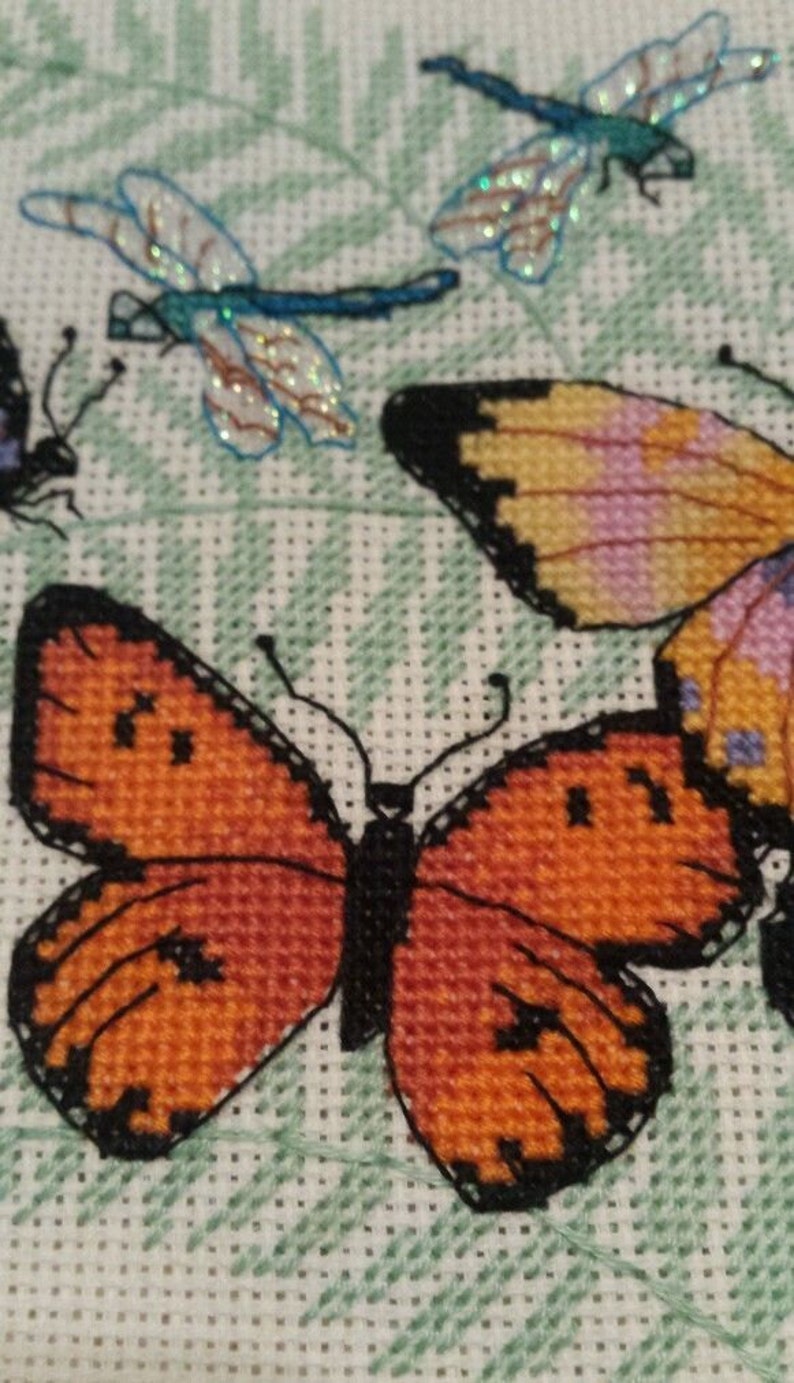K&ouml;nnte beinhalten: Kreuzstich-Stickerei mit drei Schmetterlingen. Ein Schmetterling ist orange mit schwarzen Markierungen, ein anderer ist gelb mit rosa und violetten Markierungen und der dritte ist blau mit schillernden Fl&uuml;geln.