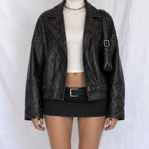 Peut inclure: Une veste en cuir noir avec des motifs floraux en relief sur les revers et les poignets, portée sur un haut blanc à col montant. La tenue comprend une mini-jupe noire avec une ceinture noire à boucle argentée et un sac à bandoulière noir.