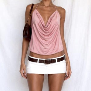 May include: A dusty pink sleeveless top with a draped cowl neckline and thin halter straps, paired with a white mini skirt featuring a dark brown leather belt with a metal buckle and a small dark brown shoulder bag hanging on the side.