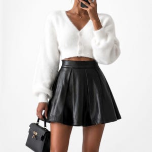 May include: A woman's outfit featuring a white, fluffy, cropped cardigan with a V-neck and button closure, paired with a black, pleated, faux leather mini skirt. A black handbag with a gold clasp is held in the hand.