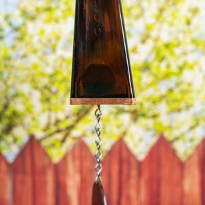 Puede incluir: Un carill&oacute;n de viento de vidrio marr&oacute;n con un borde de color cobre y una cadena plateada que cuelga de &eacute;l. El carill&oacute;n de viento est&aacute; colgado frente a una valla de madera.