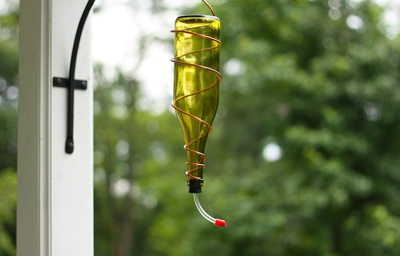 May include: A glass bottle wrapped in copper wire with a red plastic tube extending from the bottom. The bottle is hanging from a black metal hook.