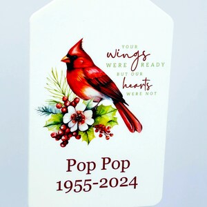 May include: A white, tag-shaped ornament featuring a vibrant red cardinal perched on a floral arrangement. Text reads "Your wings were ready but our hearts were not" and "Pop Pop 1955-2024". The ornament has a silver loop for hanging.