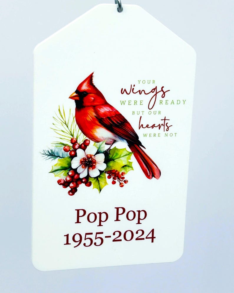 May include: A white, tag-shaped ornament featuring a vibrant red cardinal perched on a floral arrangement. Text reads "Your wings were ready but our hearts were not" and "Pop Pop 1955-2024". The ornament has a silver loop for hanging.
