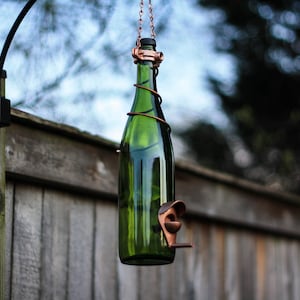 K&ouml;nnte beinhalten: Ein gr&uuml;ner Glasflaschen-Vogelfutterspender mit einem Kupferdrahtk&auml;fig und einem Kupfer-Vogelsitz. Der Spender h&auml;ngt an einem schwarzen Metallhaken, der an einem Holzzaun befestigt ist.