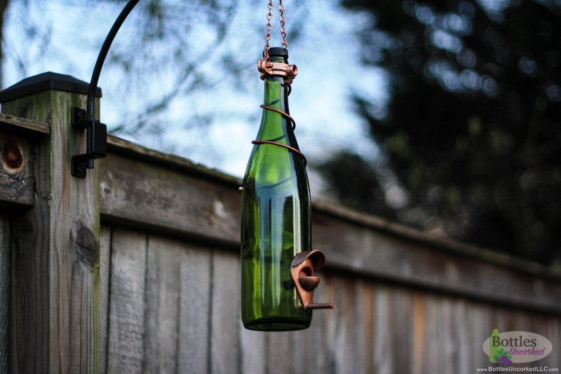 K&ouml;nnte beinhalten: Ein gr&uuml;ner Glasflaschen-Vogelfutterspender mit einem Kupferdrahtk&auml;fig und einem Kupfer-Vogelsitz. Der Spender h&auml;ngt an einem schwarzen Metallhaken, der an einem Holzzaun befestigt ist.